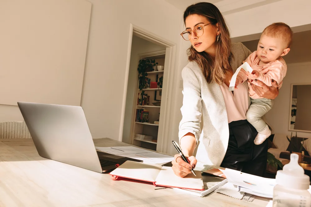 Multitasking & Work Life Balance Junge Frau im Geschäftsanzug hält ihr Baby während sie an eine Laptop arbeitet und Notize schreibt