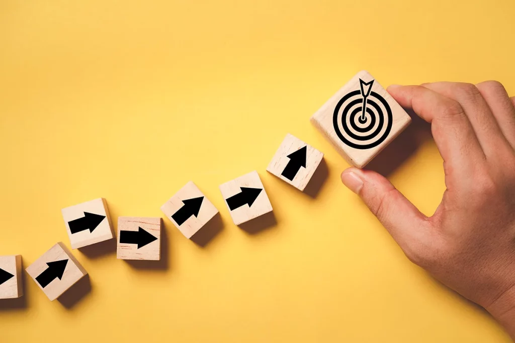 Eine Hand hält einen Holzblock mit einem Zielscheiben-Symbol, während eine Reihe von Blöcken mit Pfeilsymbolen davor liegt. Dies symbolisiert die sechs Phasen der Candidate Journey