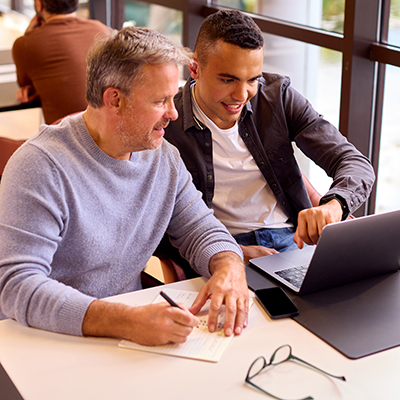 Zwei Männer sitzen an einem Schreibtisch. Der ältere Mann macht sich Notizen während der jüngere Mann ihm etwas auf einem Laptop zeigt.