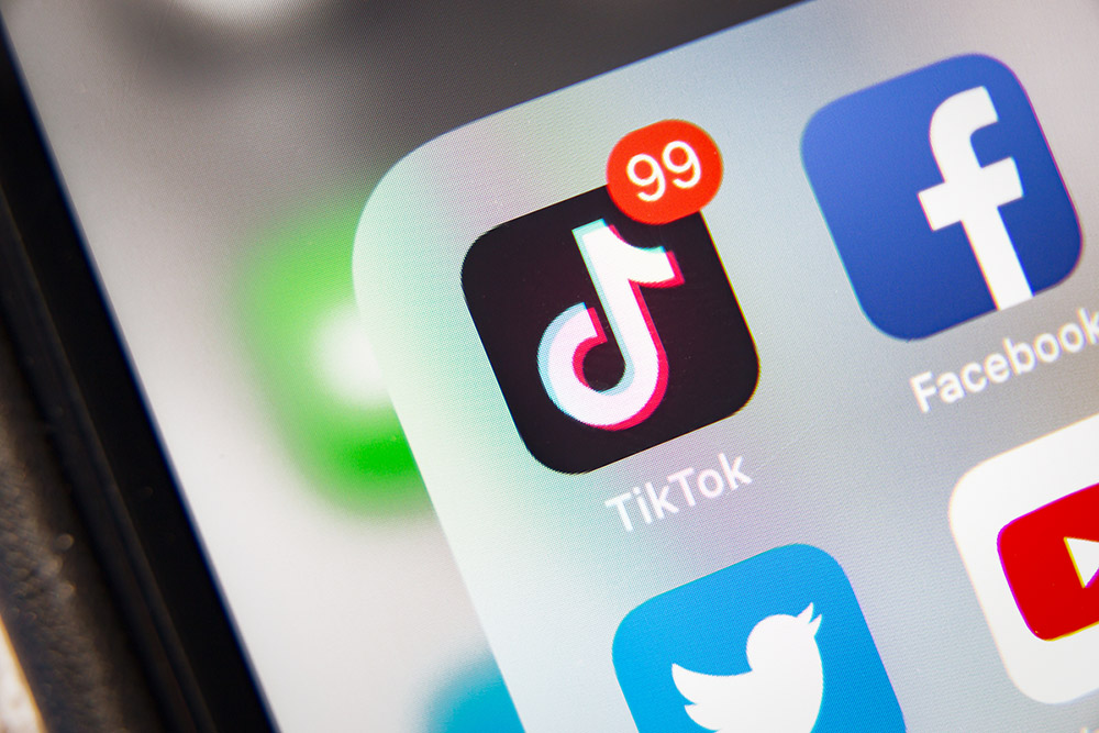 Das Bild zeigt ein Smartphone-Display mit mehreren Social-Media-Apps. Im Mittelpunkt steht das TikTok-Icon, das eine rote Benachrichtigungsmarkierung mit der Zahl 99 hat. Daneben sind Icons der Apps Facebook, Twitter und YouTube zu sehen.